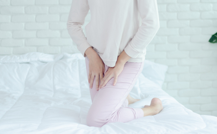 Clothed woman kneeling on bed with hands positioned in front of crotch to indicate discomfort from yeast infection.