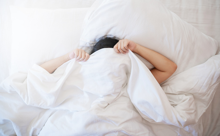 person with blanket over face lying in bed tired in the morning